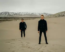Two Men Standing On A Beach With A Surfboard